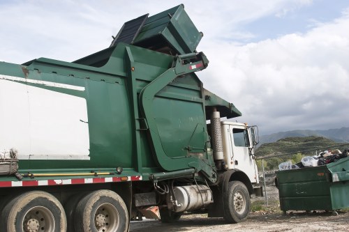 A large green industrial waste collection truck with a white cab parked outdoors on a gravel surface. The rear of the vehicle features a hydraulic lifting mechanism and an open, tilted waste hopper, indicating it is in the process of depositing or collecting rubbish. The truck's body shows signs of usage, with textured metal surfaces and some visible dirt or grime. To the right, a large green wheelie bin filled with mixed waste and debris is positioned on the ground, with some rubbish visible spilling out. The environment around the vehicle includes a paved area or driveway with distant hills and a cloudy sky overhead, suggesting a rural or semi-industrial location possibly near Kings Cross. The truck's design and context align with commercial waste removal services provided by companies like Commercial Waste Kings Cross, serving the local postcode area, promoting efficient rubbish collection and disposal in the district.