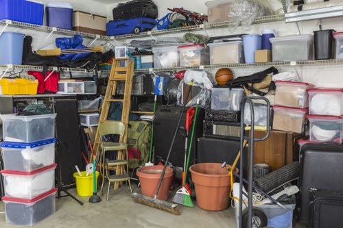 A cluttered storage area or garage in an urban setting, filled with various household and gardening items. The scene includes stacked clear plastic storage boxes with red and grey lids, some containing small items. Several garden tools such as brooms and rakes are leaning against a pile of stacked items, with a yellow bucket and mop standing among the clutter. Two terracotta plant pots are positioned in front, while a small wooden chair and a metal trolley are partially visible behind the tools. The background shows metal shelving units holding more plastic boxes, toolboxes, and miscellaneous supplies, with some items wrapped in plastic. The environment appears dimly lit, typical of a storage space used for household waste collection or clearing, representative of rubbish removal services in a city like Kings Cross. Commercial Waste Kings Cross may handle such clutter removal, emphasizing their expertise in clearing domestic and garden waste from densely stocked areas.