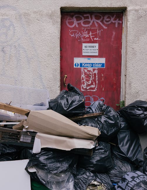 A narrow alleyway restricted space filled with a large, heavy-duty black garbage bag in the foreground, placed on a rough asphalt surface. Behind the bag, an overflowing stack of cardboard boxes, whit
