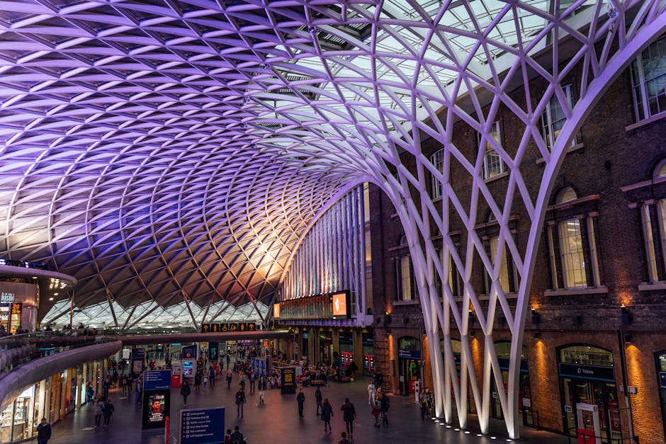 Inside a modern train station featuring a high, arched steel and glass roof structure supported by curved metal beams. The roof extends over an indoor walkway with a glass and metal railing, situated 