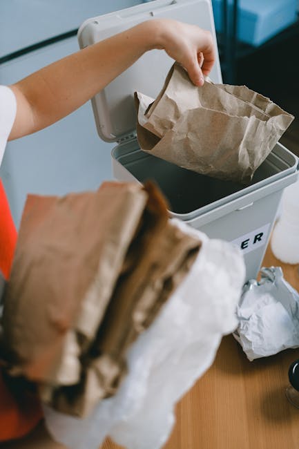 An outdoor scene showing a large collection of discarded waste materials, predominantly comprising multiple stacks of cardboard fruit and vegetable boxes, neatly piled in the center of the image. The 